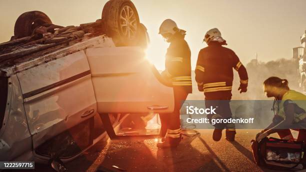 Car Crash Traffic Accident Paramedics And Firefighters Plan Rescuing Passengers Trapped In Rollover Vehicle Medics Prepare Stretchers And First Aid Equipment Firemen Use Hydraulic Cutters Spreader Stock Photo - Download Image Now