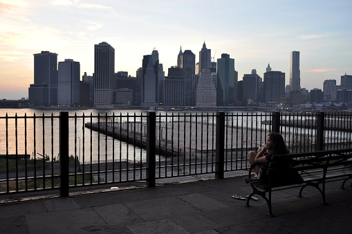A girl is sitting in a bank, enjoying the view of lower Manhattan at dusk during a summer night