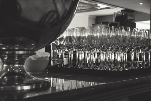 Monochrome photograph of wine glasses lined up on a bar, in preparation for a wedding reception.  View from behind the bar.