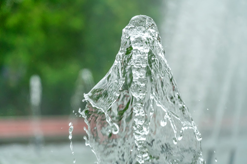 Closeup picture of a water fountain in a city park on a background of green trees