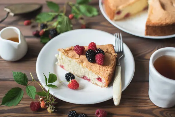 Slice of homemade sponge cake with raspberries and blackberries on a white plate on a brown wooden background