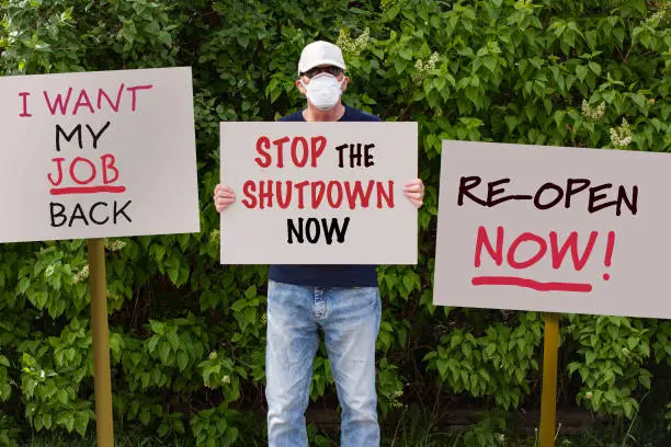 Protester with cap and medical protection mask demonstrate against stay-at-home orders due to the COVID-19 pandemic with sign saying Stop the Shutdown Now. More sign boards around him.