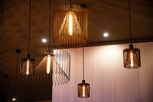 Geometric wire carcass chandeliers of different sizes. Modern metal lampshades on dark ceiling and light wall background. Lamps with glowing long light bulbs. Industrial style interior decoration