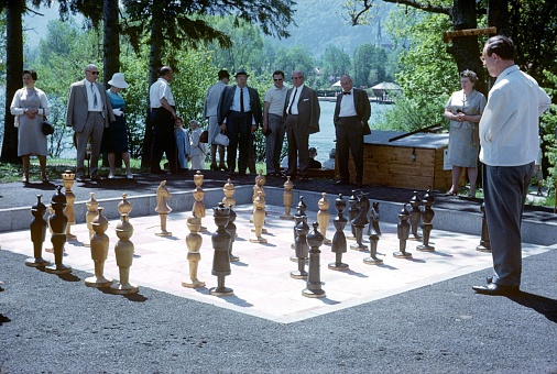 Bavaria (exact location unfortunately not known), Germany, 1966. Outdoor chess game. Two men play  chess in a community park. Spectators have gathered around the chess field.