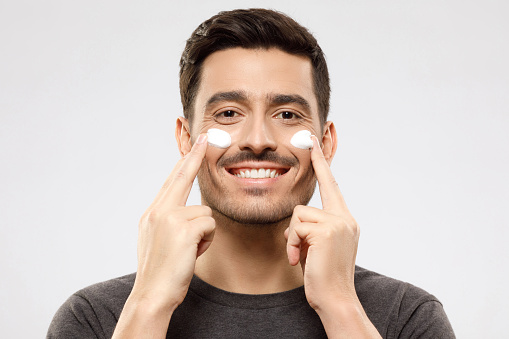 Young smiling male feeling happy about regular beauty routine, applying face cream on cheeks with fingers, feeling relaxed, isolated on gray background. Skin care men