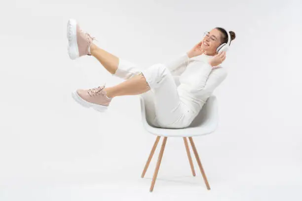 Photo of Happy woman enjoying favorite music tracks in wireless headphones, sitting on white chair, moving legs in air, feeling relaxed and happy, isolated on gray background
