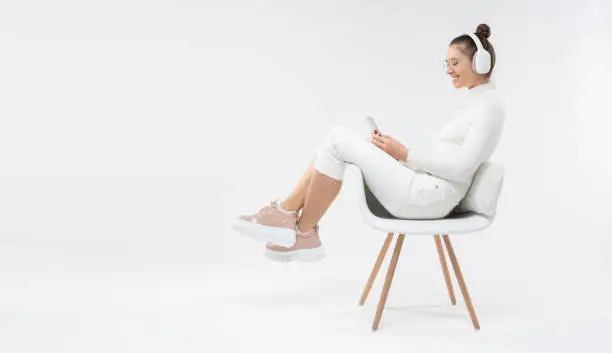 Photo of Horizontal banner of girl sitting on white chair, wearing casual clothes and trainers, listening to music in headphones, looking at phone, copy space on left, isolated on gray background