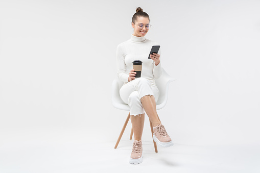 Young female sitting on white chair, checking text messages on smartphone with happy smile and holding takeaway coffee cup, isolated on gray background