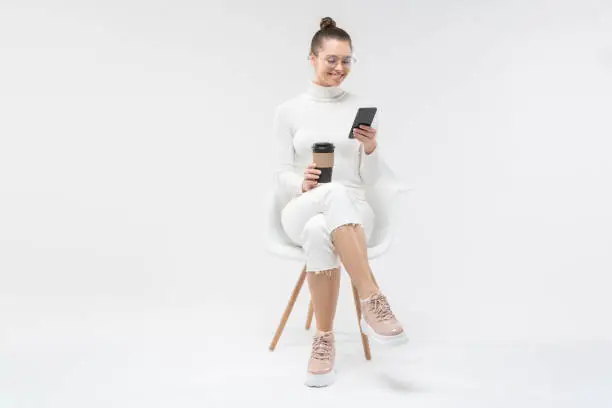 Photo of Young female sitting on white chair, checking text messages on smartphone with happy smile and holding takeaway coffee cup, isolated on gray background