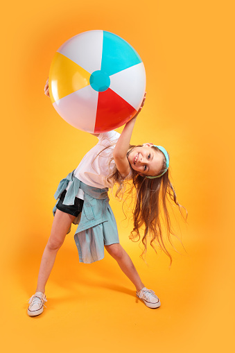 Funny happy child in summer clothes jumping with beachball on colored yellow background