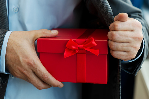 Chest view of an unrecognizable businessman wearing a suit who is about to give a gift box out of his jacket. The red gift box is wrapped with red ribbon.
