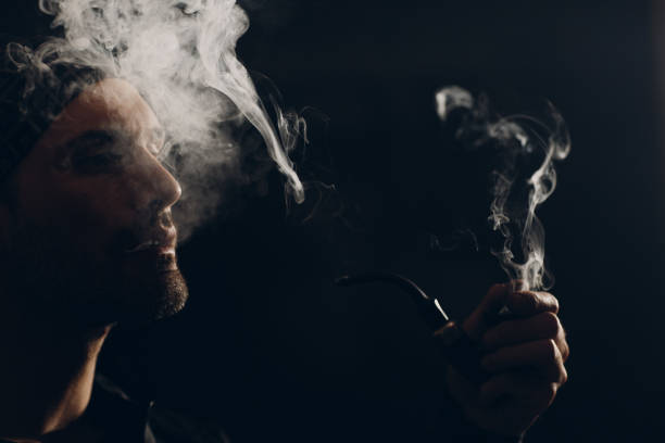 istock Man smoking a pipe on dark background. Profile portrait with clouds of smoke and back light. 1222651200