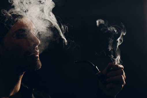 Man smoking a pipe on dark background. Profile portrait with clouds of smoke and back light.