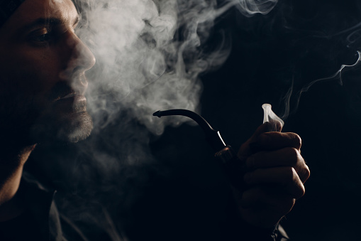 Man smoking a pipe on dark background. Profile portrait with clouds of smoke.