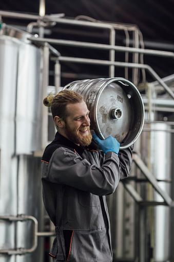 Happy man working at a brewery carrying beer aging barrels.