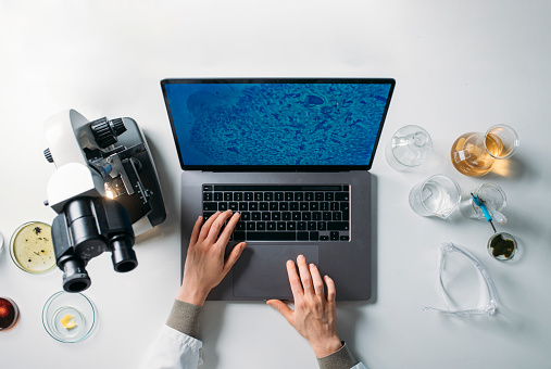 Scientist using a laptop computer along with a microscope and other research-related objects.
