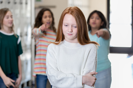 Sad elementary age girl standing with arms crossed and frowning while other student point and laugh at her