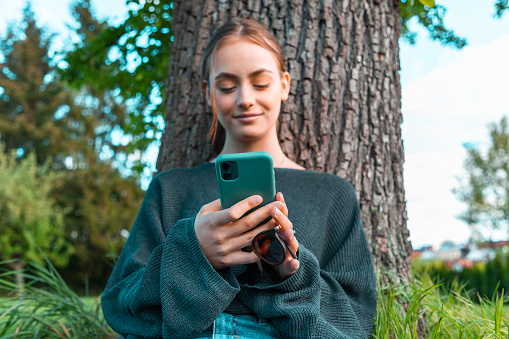 Confident smiling teenage woman leaning on tree in the garden, reading messages on her mobile phone, smiling confident and happy. Young women social media lifestyle concept shot.