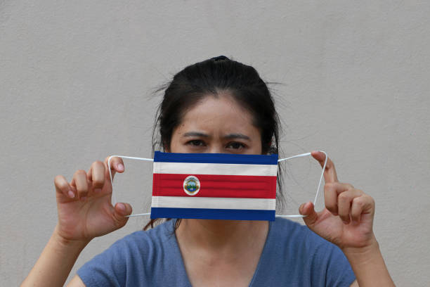 une femme avec le drapeau du costa rica sur le masque hygiénique dans sa main et a soulevé le visage avant sur le fond de couleur beige. tiny particle ou virus corona ou covid 19 protection. - arms lifted photos et images de collection