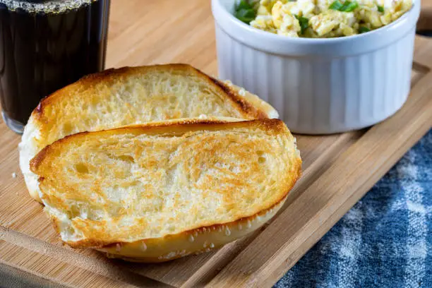 Photo of traditional Brazilian breakfast. Coffee in a glass, toasted bread and butter, and scrambled eggs, on top of a wooden board and a blue cloth (Pão na Chapa)