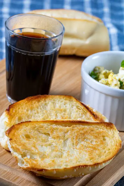 Photo of traditional Brazilian breakfast. Coffee in a glass, toasted bread and butter, and scrambled eggs, on top of a wooden board and a blue cloth (Pão na Chapa)