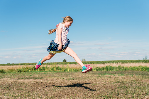 A smilling little girl is jumping in the air in a countryside. She has a fun expression. She looks free and happy. She has long tied hair. She is wearing a blue skirt and a pink shirt. She is in Quebec, Canada.