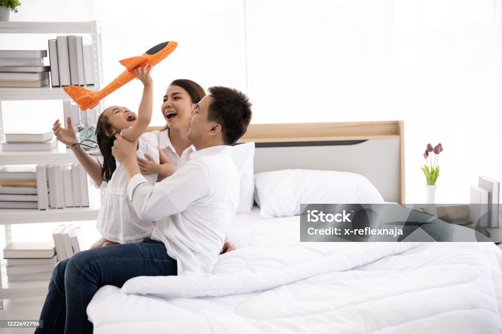Asian family happy on the bed in the house with sunlight from the window. Father, mother and daughter, aged three are playing with a plane together. Family leisure activities Asian and Indian Ethnicities Stock Photo