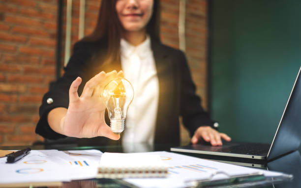 istock a businesswoman holding a glowing light bulb while working on laptop computer and paperwork in office 1222701501