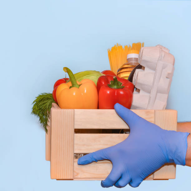 Hands in medical gloves gives set of food in a wooden box on a blue background.Coronavirus Relief Funds and Donations.Сoncept of safety mail goods courier delivery.The concept of online orders manos en guantes médicos da conjunto de alimentos en una caja de madera sobre un fondo azul. fondos de socorro de coronavirus y donaciones.oncept de la entrega de mensajería de mercancías de correo de seguridad. el concepto de pedidos en línea