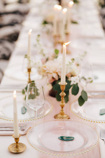 istock Wedding dinner table at reception. Beautiful white delicate candles burn in metal candlesticks, against background of white and pink tablecloth, flower arrangements and glass plates with gold beads 1222720033