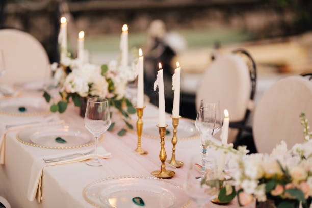 istock Wedding dinner table at reception. Beautiful white delicate candles burn in metal candlesticks, against background of white and pink tablecloth, flower arrangements and glass plates with gold beads 1222720210