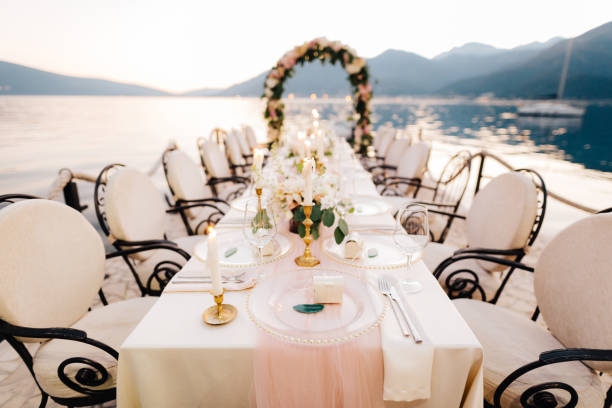 istock Close-up of a wedding dinner table reception. A table stands on beach overlooking mountains at sunset. Metal forged chairs, burning candles, wedding arch of flowers, cream-colored cloth tablecloth. 1222721996
