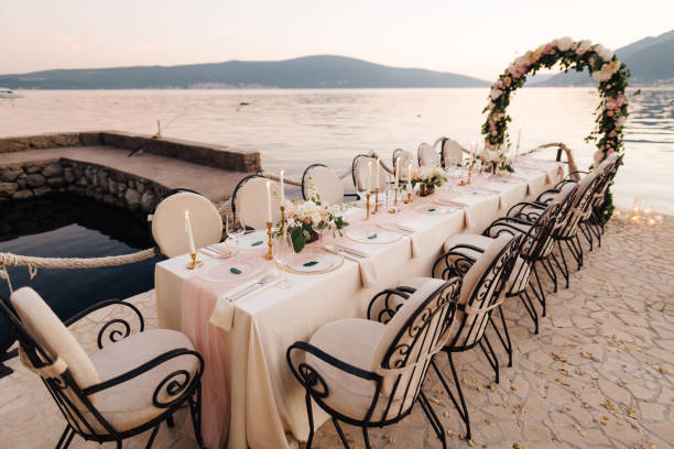 istock Close-up of a wedding dinner table reception. A table stands on beach overlooking mountains at sunset. Metal forged chairs, burning candles, wedding arch of flowers, cream-colored cloth tablecloth. 1222722336