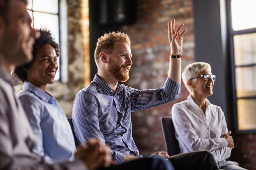 Happy business people sitting on an education event in a board room. Focus is on redhead man raising his hand.