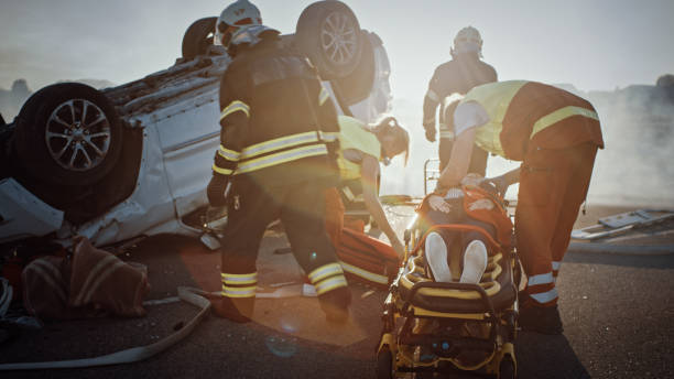 sulla scena dell'incidente stradale: la squadra di soccorso dei vigili del fuoco tira fuori la vittima dal veicolo di ribaltamento, usano le barelle con attenzione, la consegnano ai paramedici che eseguono il primo soccorso - car fire accident land vehicle foto e immagini stock