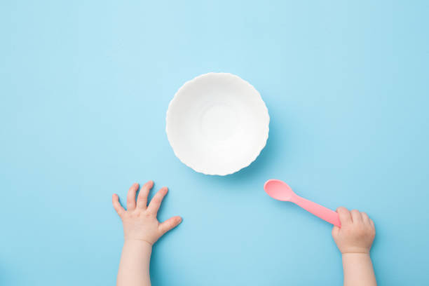 mains de chéri retenant la cuillère en plastique rose et attendant la nourriture. bol blanc vide sur le fond bleu clair de table. couleur pastel. closeup. tir de point de vue. vue en haut vers le bas. - baby food photos et images de collection