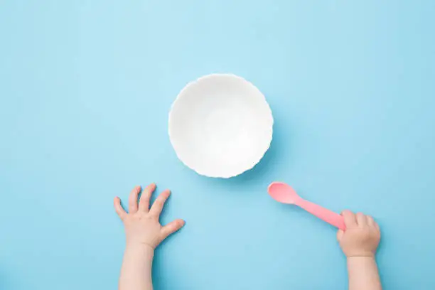 Baby hands holding pink plastic spoon and waiting food. Empty white bowl on light blue table background. Pastel color. Closeup. Point of view shot. Top down view.