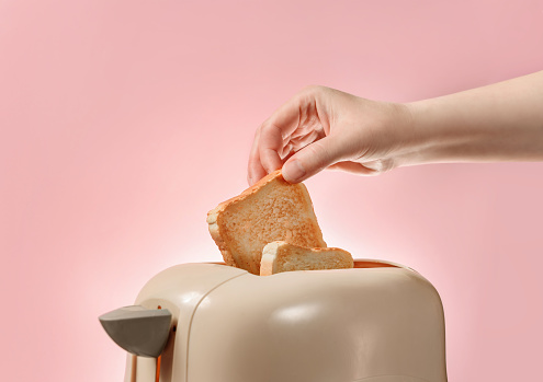 A female hand takes out toasted bread toast from a toaster. Toaster with ready-made toasts and a female hand isolated on a pink background with a copy space.