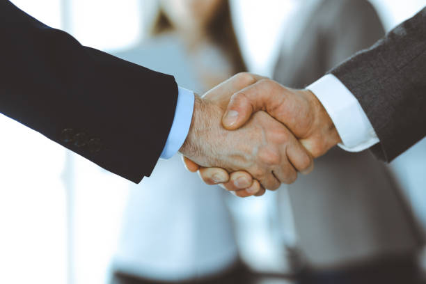 istock Business people shaking hands while standing with colleagues after meeting or negotiation, close-up. Group of unknown businessmen and women in modern office. Teamwork, partnership and handshake concept, toned picture 1222730639