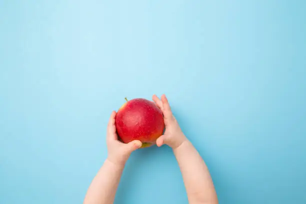 Baby hands holding red apple. Fresh fruit. Empty place for text on light blue table background. Pastel color. Closeup. Point of view shot. Top down view.