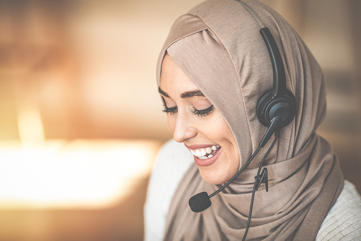 Woman wearing headscarf and headset talking while working in a call centre office, smiling, close-up. Close up beautiful muslim woman call center agent wear headset device and smiling working in operation room