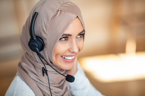 Portrait of a friendly young muslim call center agent working in a modern office. Portrait of beautiful arabic woman in hijab smiling and looking at camera while working in office with copy space.