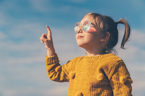 Five years little girl with ponytails, dressed in a yellow sweater and funny sunglasses is looking to the blue sky.