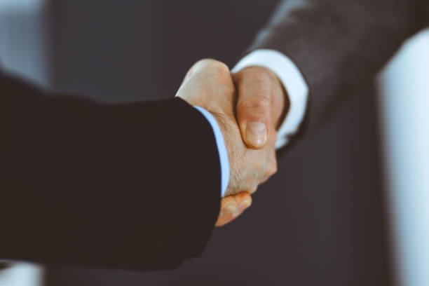 istock Business people shaking hands while standing with colleagues after meeting or negotiation, close-up. Group of unknown businessmen and women in modern office. Teamwork, partnership and handshake concept, toned picture 1222746129