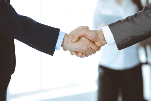 istock Business people shaking hands while standing with colleagues after meeting or negotiation, close-up. Group of unknown businessmen and women in modern office. Teamwork, partnership and handshake concept, toned picture 1222746139