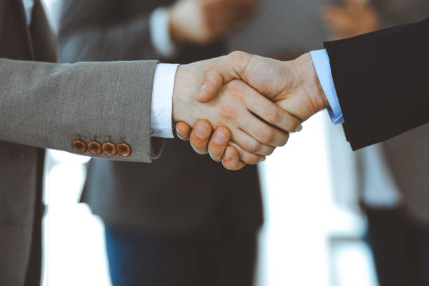 istock Business people shaking hands while standing with colleagues after meeting or negotiation, close-up. Group of unknown businessmen and women in modern office. Teamwork, partnership and handshake concept, toned picture 1222746152