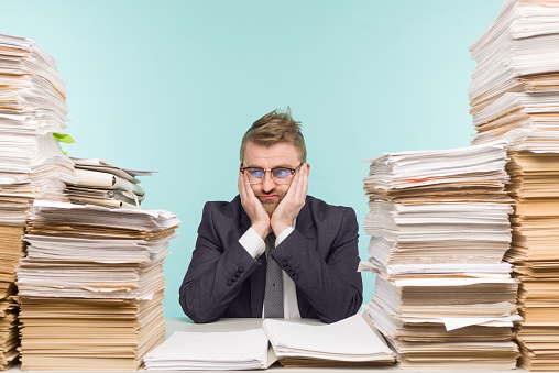 Shocked businessman sitting at the table with many papers in office, he is overloaded with work - image