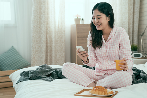 carefree young girl in pajamas eating breakfast in bed while using mobile phone. relax cute asian korean woman holding glass of fresh orange juice in morning bedroom. sunshine through window.