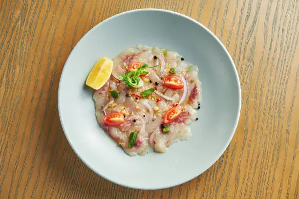 Chilled sea bass carpaccio with red onion, cherry tomatoes and hot chili peppers in a beige bowl on a wooden background. Close up, selective focus