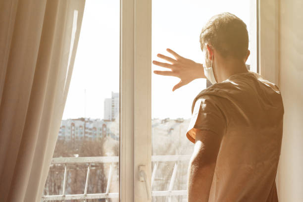 istock Student freelancer with computer for distance job due to coronavirus quarantine. Young man in medical protective mask on face sitting on windowsill and using laptop. Hipster blogger working at home 1222763572